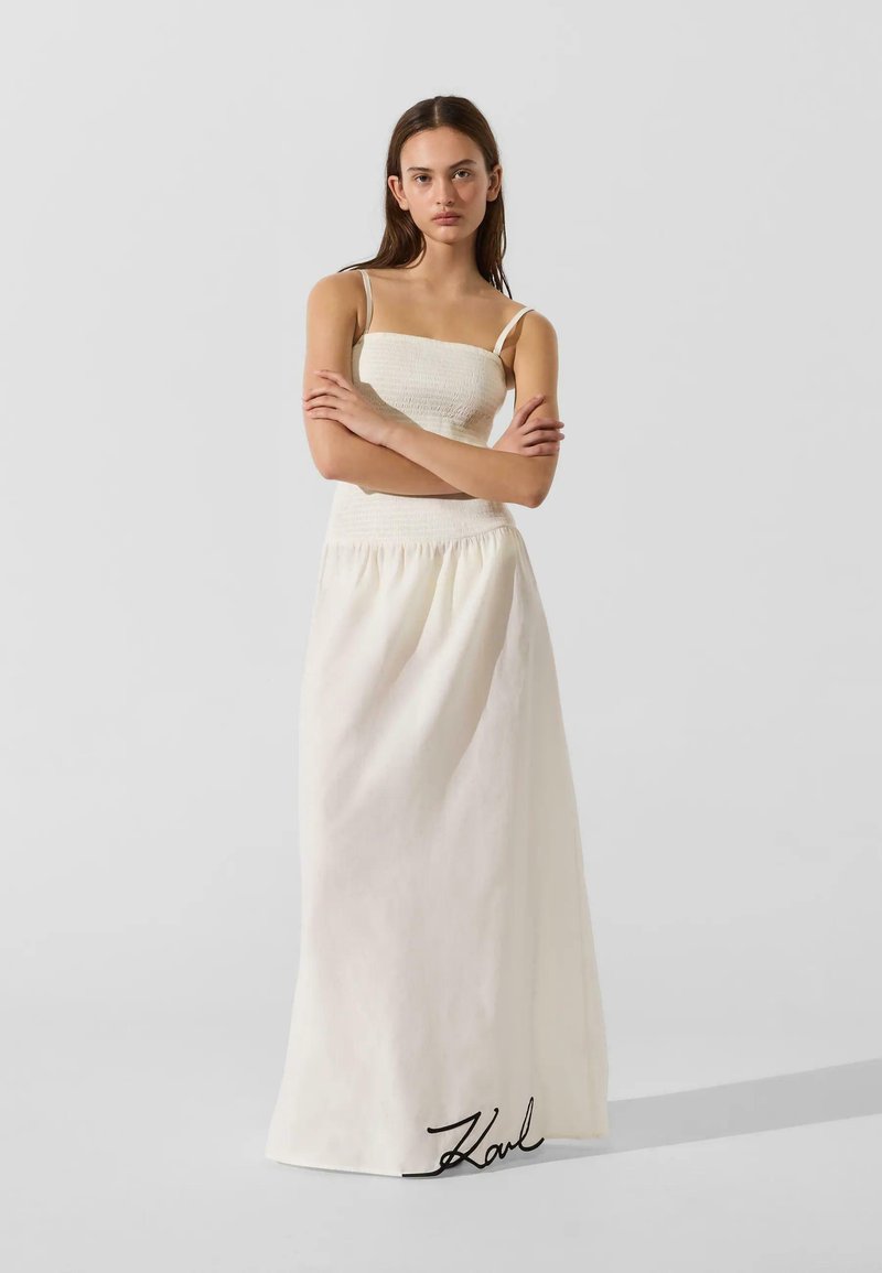 Femme aux longs cheveux foncés portant une robe maxi blanche sans manches avec un corsage smocké, debout les bras croisés devant un fond clair uni.
