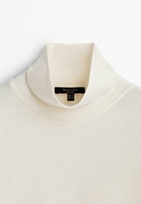 Cremfarbener Rollkragenpullover aus weicher Baumwolle, mit einem weiten Kragen, nahtlosem Design und einem dezenten Glanz. Etiketteninformationen sind sichtbar innen.