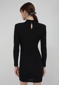 Schwarz, langärmliges Kleid in taillierter Form, mit hohem Kragen und einer Aussparung am Rücken. Glatte Textur mit leichtem Schimmer.