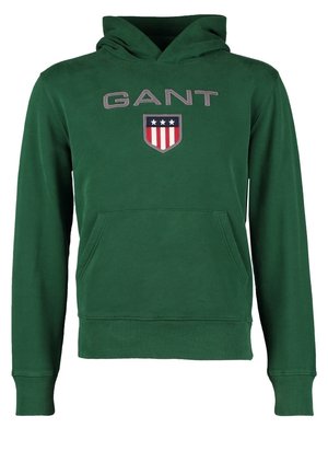 Mand gående iført en grøn GANT hættetrøje, olivengrønne cargobukser, sorte og hvide sneakers, mod en ensfarvet lys baggrund.