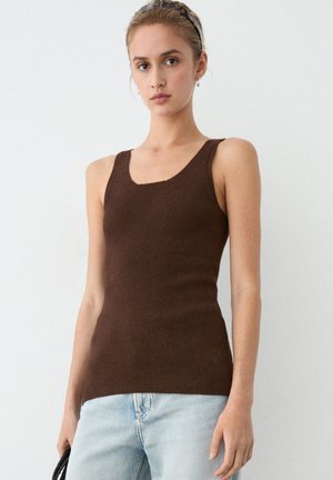 Jeune femme portant un débardeur côtelé marron et un jean bleu clair, tenant un sac à main noir sur un fond blanc uni.