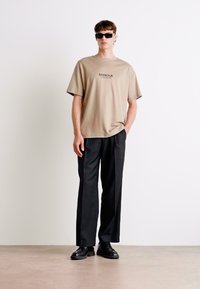 Beez värvi puuvillane t-särk, millel on "BARBOUR" logo, mida on kombineeritud mustade laia lõikega pükstega ja mustade kingadega. Mudel kannab tumedat päikeseprille.