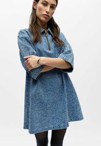 Femme aux longs cheveux bruns portant une robe bleue texturée à manches courtes avec une fermeture éclair frontale, des collants noirs et un bracelet argenté, posant les bras croisés.