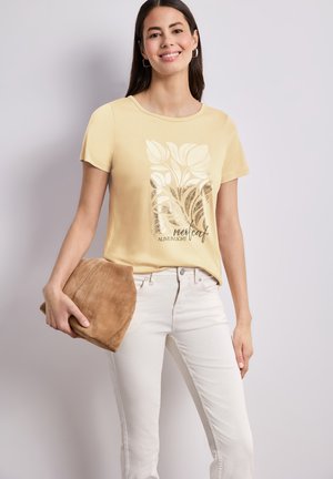 Kobieta nosząca żółtą koszulkę z krótkim rękawem z botanicznym nadrukiem i napisem, trzymająca beżową zamszową kopertówkę, zestawiona z kremowymi spodniami.