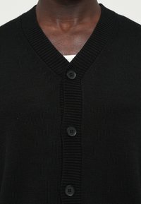 Dėtis iš arti, kur asmuo dėvi juodą v-neck megztinį su užsagomis virš baltos marškinės, matosi kaklo ir viršutinės krūtinės sritis.