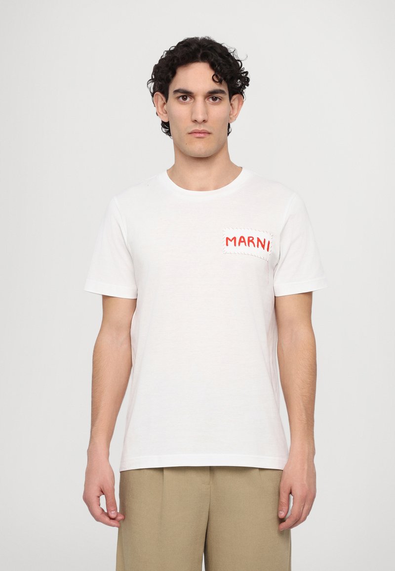 White cotton t-shirt featuring a red embroidered logo patch on the left chest and a classic crew neck design. Shorts in beige.