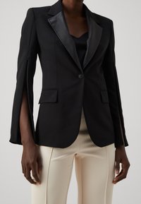 Czarna marynarka z satynowym klapą, dopasowany krój i otwartym rękawem. Posiada dwie przednie kieszenie i pojedyncze zapięcie na guzik.