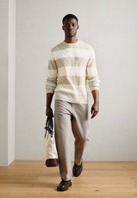 Helles beiges Sweatshirt mit horizontalen Streifen, hellgraue Hose und dunkelbraune Slipper. Trägt eine weiße Tasche mit braunem Akzent.
