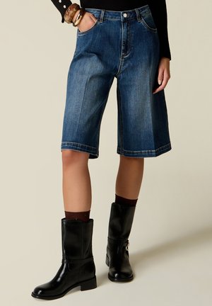 Vrouw die knielange blauwe denimshorts draagt, zwarte midkuitlaarzen en een zwarte top met lange mouwen, staat met één hand in de zak.
