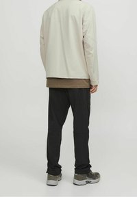 Giacca a maniche lunghe beige chiaro sopra una camicia marrone, abbinata a pantaloni cargo neri e sneakers grigie. Tessuto morbido con design minimalista.