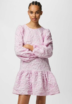 Ung kvinne med fletter, iført en langermet lys rosa teksturert kjole, stående med armene i kors foran en ensfarget bakgrunn.