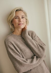 Beige übergroßer Strickpullover mit Rippenstruktur und langen Ärmeln, lässig getragen mit einer Hand am Hals. Schlichtes Design, weicher Stoff.