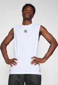 T-shirt de sport sans manches blanche avec un logo noir sur la poitrine, en matériau lisse. Porté avec un short noir texturé, les mains sur les hanches.