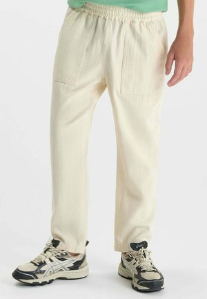 Persoon draagt crèmekleurige broek met elastische taille en voorzakken en zwart-witte hardloopschoenen, staand tegen een witte achtergrond.