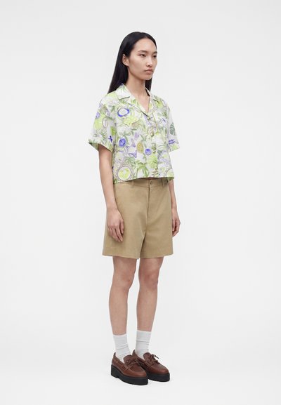 Νεαρή γυναίκα στέκεται φορώντας μια κοντομάνικη φλοράλ πουκάμισο με κουμπιά, μπεζ σορτς μέχρι το γόνατο, λευκές κάλτσες και καφέ παπούτσια με πλατφόρμα.