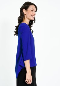 Top blu royal con maniche a tre quarti, caratterizzato da una vestibilità rilassata e un orlo posteriore allungato. Tessuto morbido; indossato con pantaloni neri.