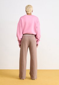 Rosa oversized sweatshirt med sänkta axlar, i kombination med lösa, bruna, medelhöga sweatpants. Släta texturer mot ett gult golv.