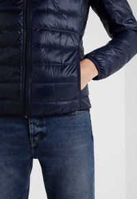 Námorně modrá zateplená bunda se zipem vpředu, bočními kapsami a elastickými manžetami. Hladká, lesklá textura a vodorovné švy.