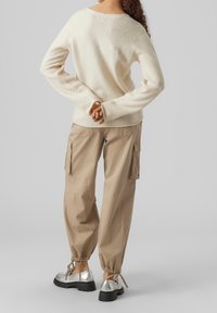 Kremowy sweter z dzianiny o luźnym kroju, beżowe spodnie typu cargo z bocznymi kieszeniami oraz błyszczące srebrne buty na sznurowadła z czarnymi podeszwami.