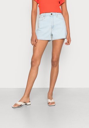 Pantalones cortos de mezclilla azul claro de cintura alta, con cuatro bolsillos y costuras blancas. Combinados con una blusa de canalé naranja brillante y sandalias blancas.
