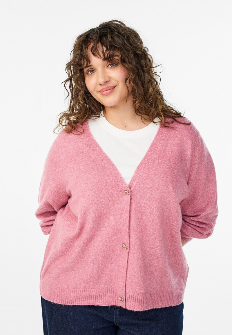 Rozā adīta džemperis ar V-veida kakla izgriezumu, ar trim pogām, ribota apakšmala un garām piedurknēm. Valkāts pāri baltai krekliņai.