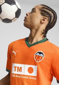 Oranžový futbalový dres sTmavozeleným golierom a logom, textúrovaná látka, obsahuje text sponzora a biele logo Puma na boku. Čierno-biely futbalový lopta nad ním.