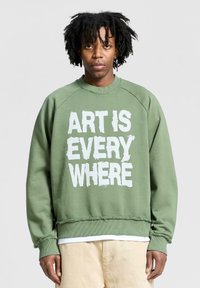 Felpa verde con collo a giro, maniche raglan e orlo sfrangiato. Testo "L'ARTE È OVUNQUE" in bianco, carattere in grassetto sul fronte.