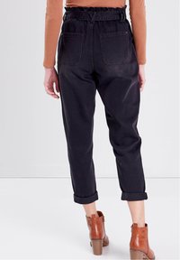 Person som bär svarta högmidjade papperssäckbyxor med uppvikt fåll, bruna klackboots och en långärmad brun topp, står på ett vitt golv.