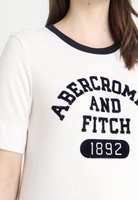 Bílý tričko s krátkým rukávem a námořnický ribovaný výstřih, s černě vyšitým textem: "Abercrombie and Fitch 1892" na přední straně.