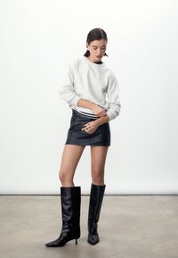 Grijze trui, zwarte leren minirok en zwarte kniehoge stiletto laarzen. Gladde texturen, nauwsluitend ontwerp en effen kleurenpalet.