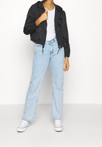 Svart kort jacka med huva, dragsko och dragkedjor; bärs över en vit skjorta och kombineras med ljusblå raka jeans och vita sneakers.