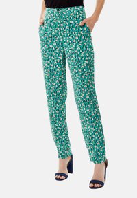 Green patterned trousers with a geometric design in pink and blue. Features pocket details and a relaxed fit, paired with navy heeled sandals.
