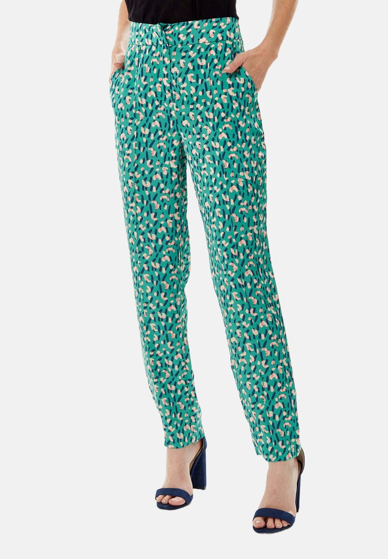 Green patterned trousers with a geometric design in pink and blue. Features pocket details and a relaxed fit, paired with navy heeled sandals.
