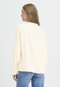 Long-sleeve, cream-colored shirt made from smooth fabric, featuring a back yoke and relaxed fit. Paired with light blue jeans.