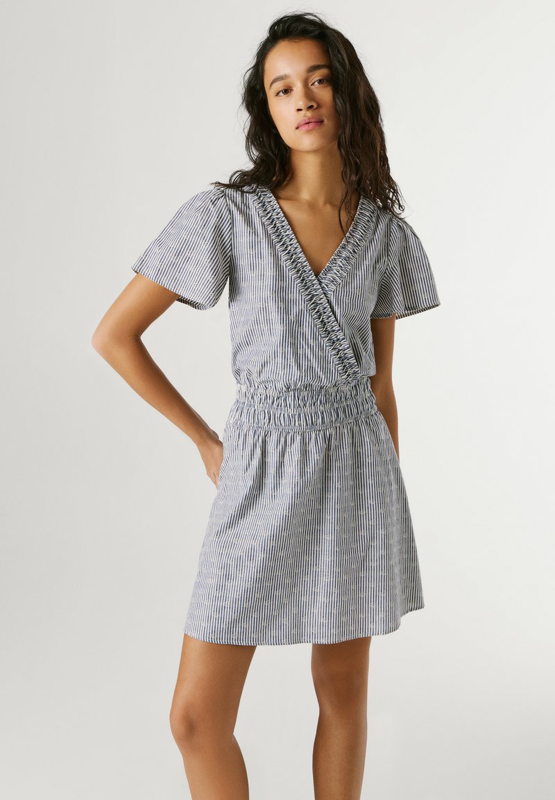 Mlada žena u prugastoj haljini kratkih rukava do koljena s V-izrezom i skupljenim strukom, stoji s jednom rukom na kukovima.