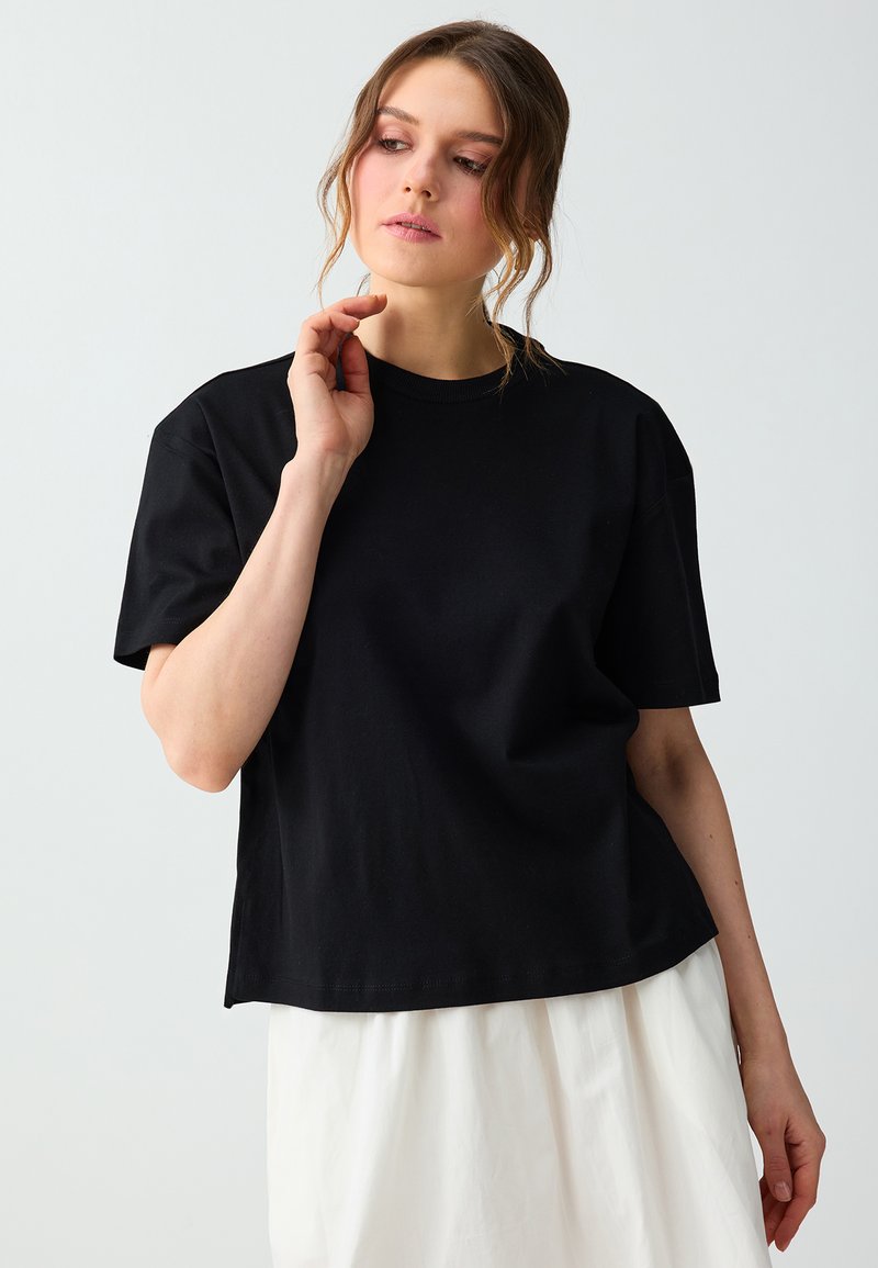 Femme à la peau claire et aux cheveux bruns ondulés, portant un t-shirt noir à manches courtes et une jupe blanche, regardant vers le bas avec la main près du visage.