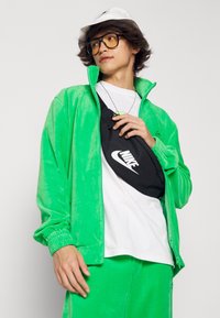 Grön velour träningsoveralljacka och byxor, vit T-shirt, svart Nike midjeväska, vit keps; har minimala logotyper och en avslappnad silhuett.