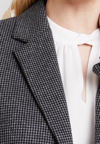 Blazer a motivo houndstooth grigio con revers a punta sopra una camicetta bianca con un piccolo apertura a chiave. Vestibilità su misura e tessuto testurizzato.