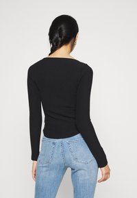 Kvinde med fletning iført en tætsiddende sort langærmet trøje og lyseblå højtaljede jeans, set fra ryggen mod en ensfarvet baggrund.