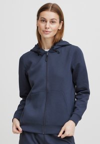 Felpa con zip color navy, dotata di cappuccio regolabile con cordino, tasche frontali e polsini a costine. Presenta un piccolo logo sul lato sinistro del petto. Tessuto morbido.