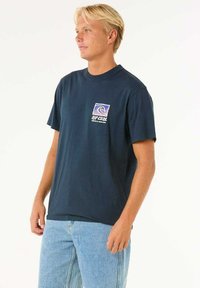 Navy cotton t-shirt with crew neck, short sleeves, and a front pocket featuring a colorful logo graphic. Worn with light denim shorts.