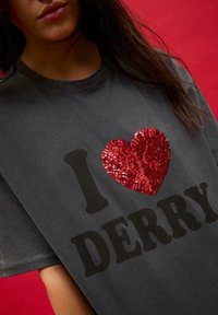 Szara koszulka z okrągłym dekoltem, z dużym szkarłatnym sercem wyszywanym cekinami oraz wyraźnym czarnym napisem "I DERRY". Materiał wygląda na miękki i lekko zniszczony.
