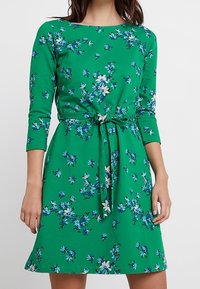 Zelené šaty s motivem vzoru, tříčtvrteční rukávy, květinový design v modré a bílé, přiléhavý pas se zavazováním ve stejné barvě, měkká tkanina.