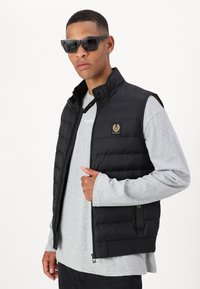 Black puffer vest with a high collar, zip closure, and logo patch. Worn over a light gray long-sleeve shirt, model wearing sunglasses.