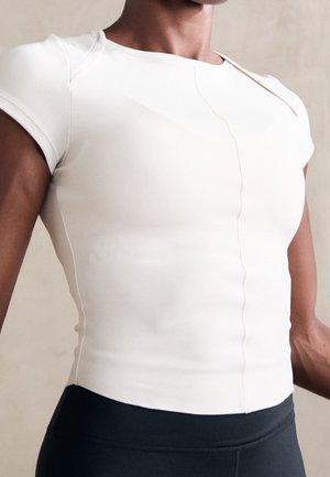 Vrouw die een nauwsluitend wit shirt met korte mouwen draagt, gecombineerd met een zwarte broek met hoge taille, tegen een neutrale achtergrond.