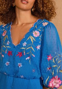 Blå broderad klänning med V-halsringning, scallopade kanter, puffärmar och intrikata blommönster i rosa och vitt. Strukturerad tyg.