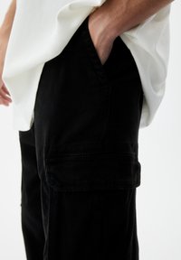 Zwarte cargobroek met een relaxed fit, voorzien van zijzakken en een gladde textuur. Gecombineerd met een witte, iets losse t-shirt.