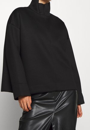 Zwart pullover met een hoge kraag, gemaakt van zachte stof. Heeft verlaagde schouders en een losse pasvorm, gecombineerd met een glanzende zwarte leren rok.