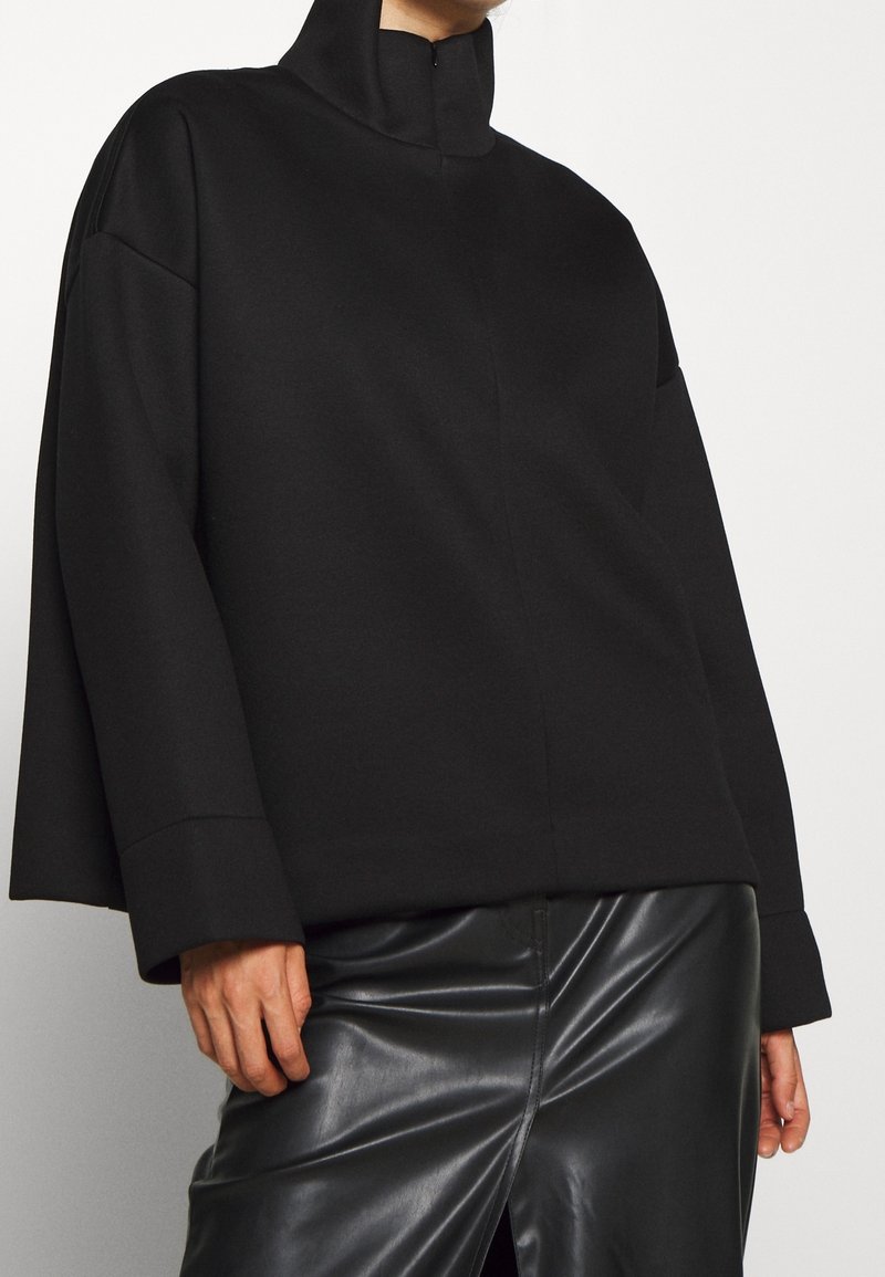 Černý pulovr s vysokým límcem, vyrobený z měkké látky. Má spadlé ramena a volný střih, je kombinován s lesklou černou koženou sukní.