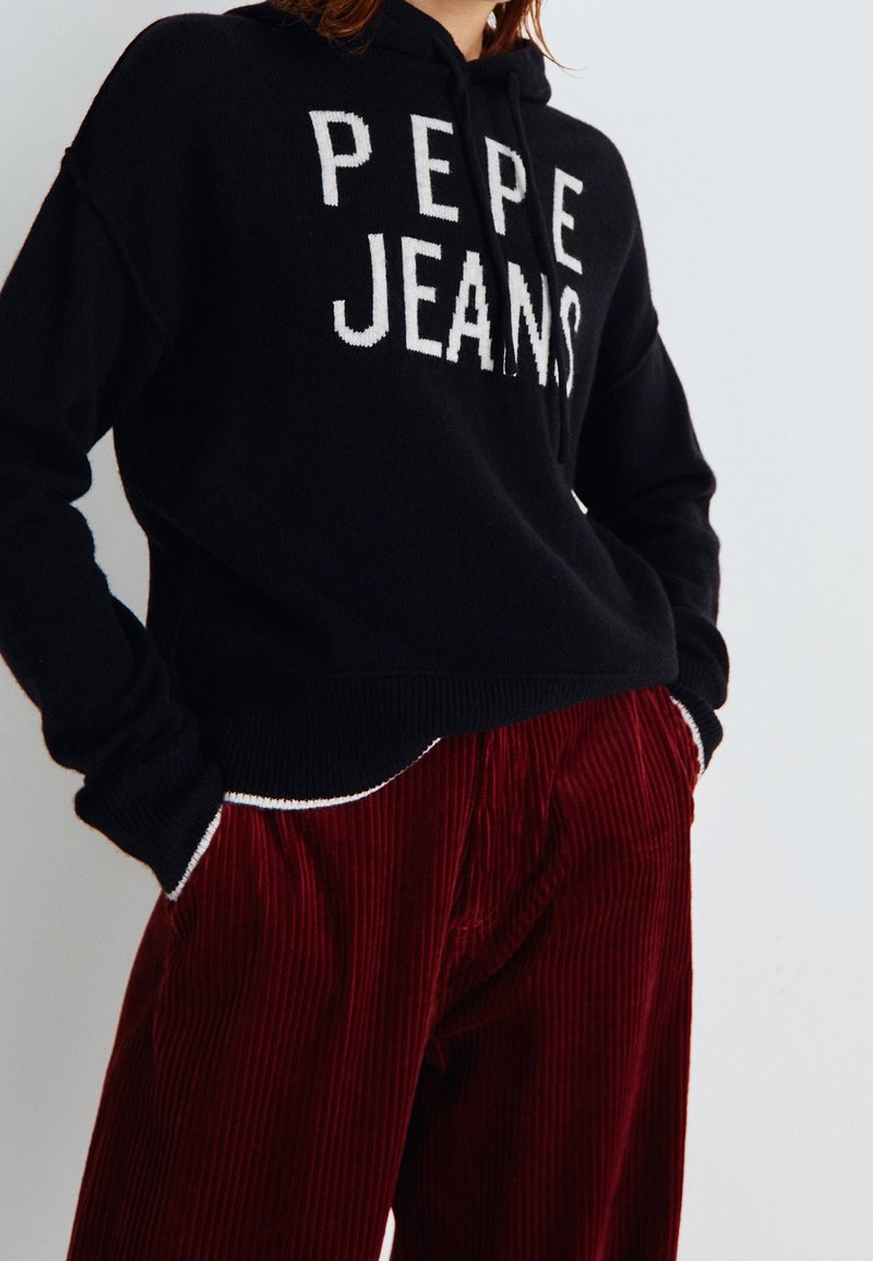 Sweat à capuche noir avec le logo "PEPE JEANS" en texte blanc. Associé à un pantalon en velours côtelé rouge avec des nervures verticales et des accents de bordure blanche.